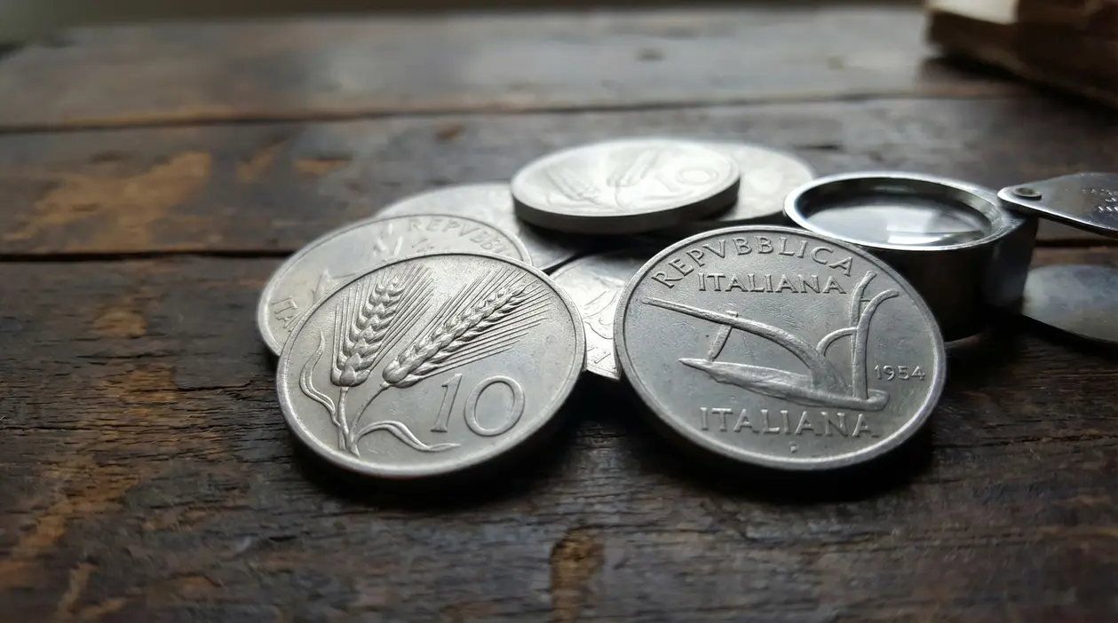 Monete da 10 lire con la spiga su un tavolo di legno, accanto a una lente per il controllo numismatico